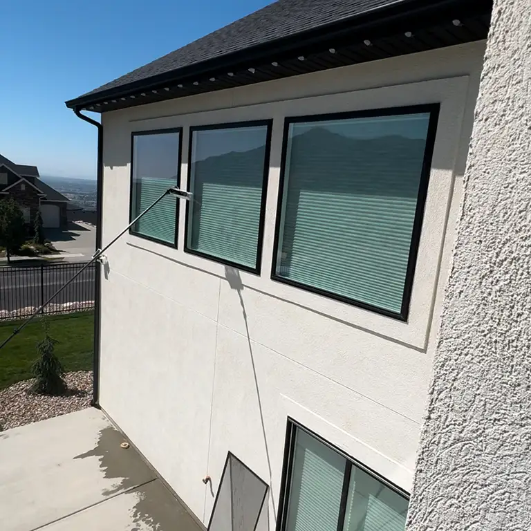 A water-fed pole with a brush cleans second-story windows on a beige house under a clear sky.