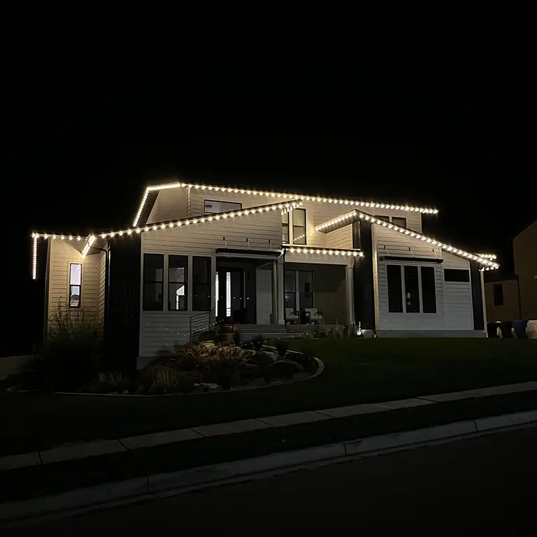 Modern house outlined with warm white lights, illuminating the roofline and front yard at night.