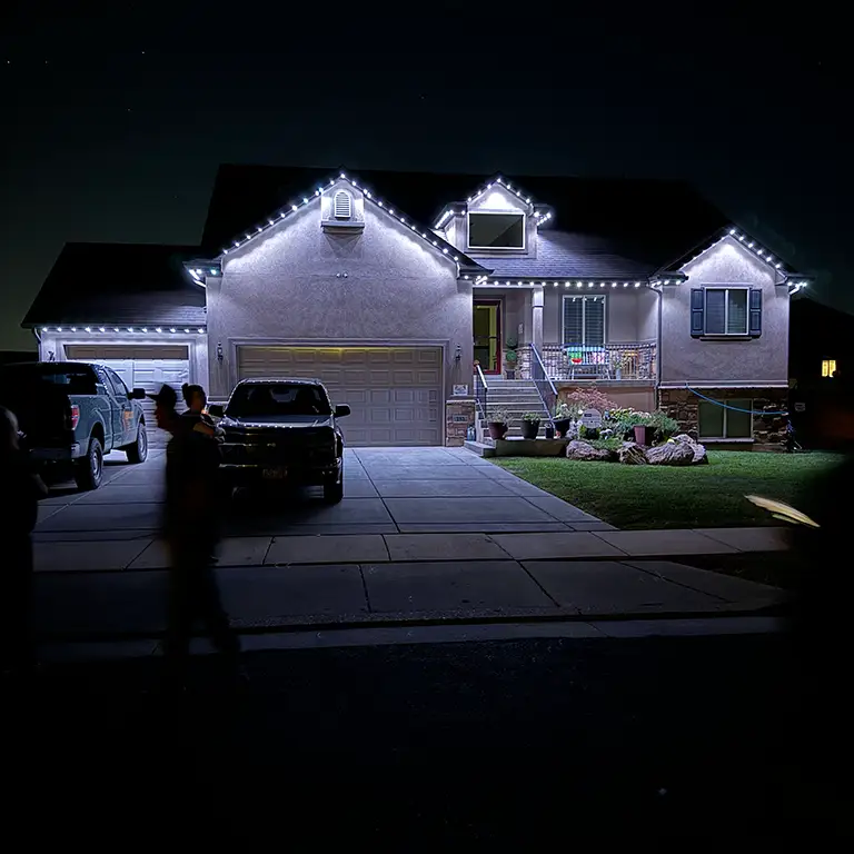 House at night with bright white string lights outlining the roof and a lit driveway with parked cars.