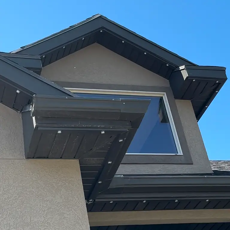 Close-up of a gable roof with black trim, featuring a large triangular window under a clear blue sky.