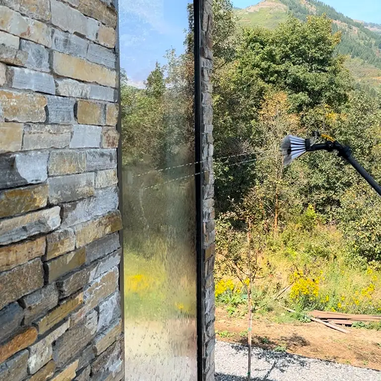 Brush tool spraying water to clean a glass window on a stone building with trees and hills in the background.