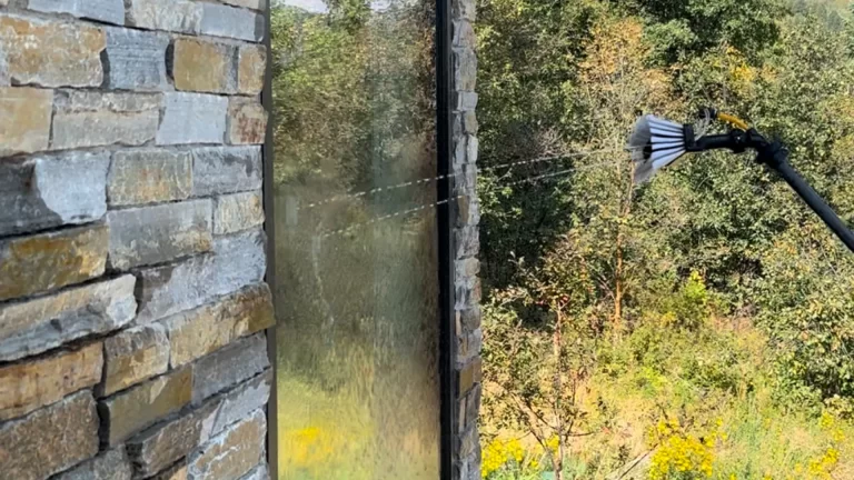 A brush on a pole sprays water while cleaning a tall window framed by a stone wall with trees in the background.