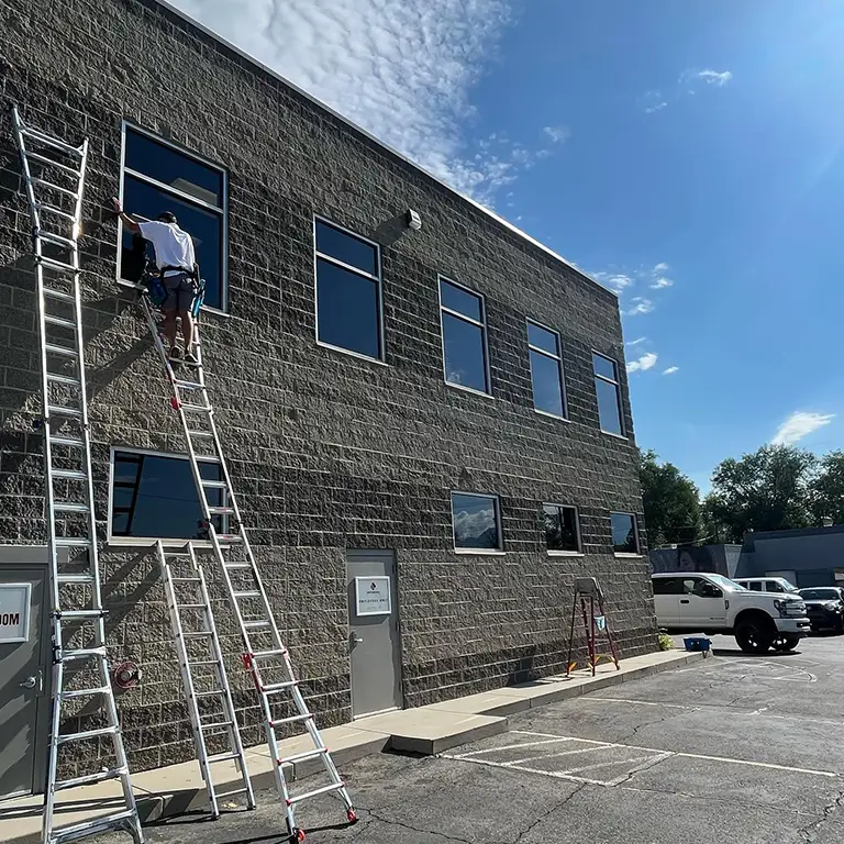 Worker cleaning second-story windows of a brick building while standing on a ladder, with multiple ladders and tools visible in the parking lot below.