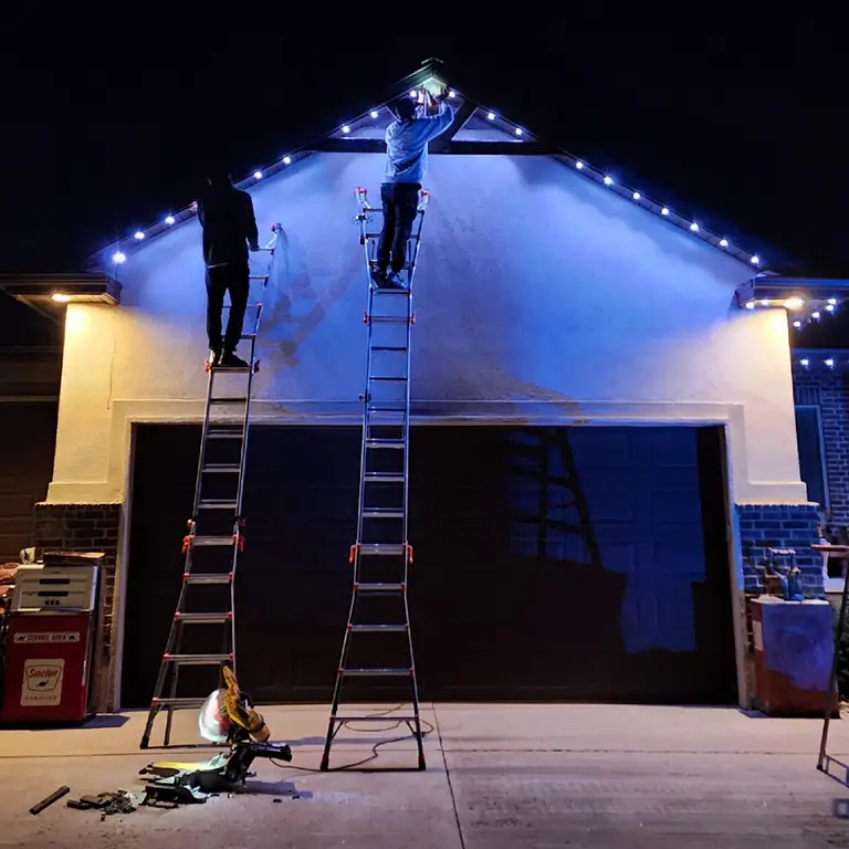 Two workers on ladders installing holiday lights along the roofline of a house at night, with tools and equipment on the driveway.