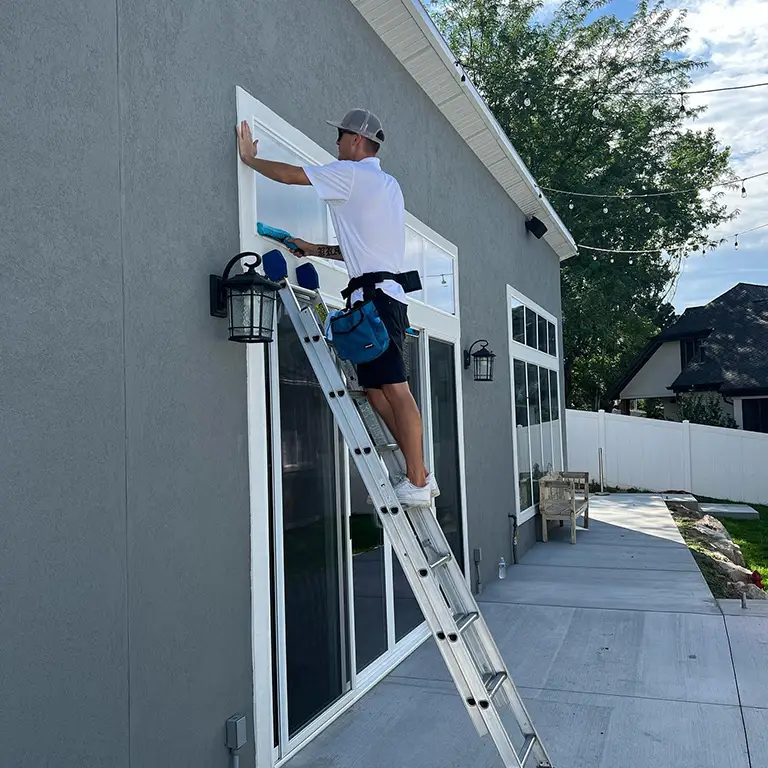 Worker on a ladder cleaning a large window of a residential home, with cleaning tools attached to their belt.