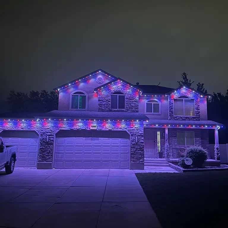 House with red and blue holiday lights outlining the roof and windows, lit at night.