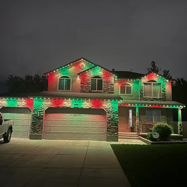 House decorated with red and green holiday lights, illuminating the driveway and stone facade at night.