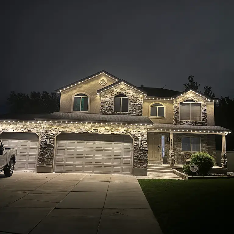 Two-story house with stone accents, lit by warm white string lights along the roofline at night.