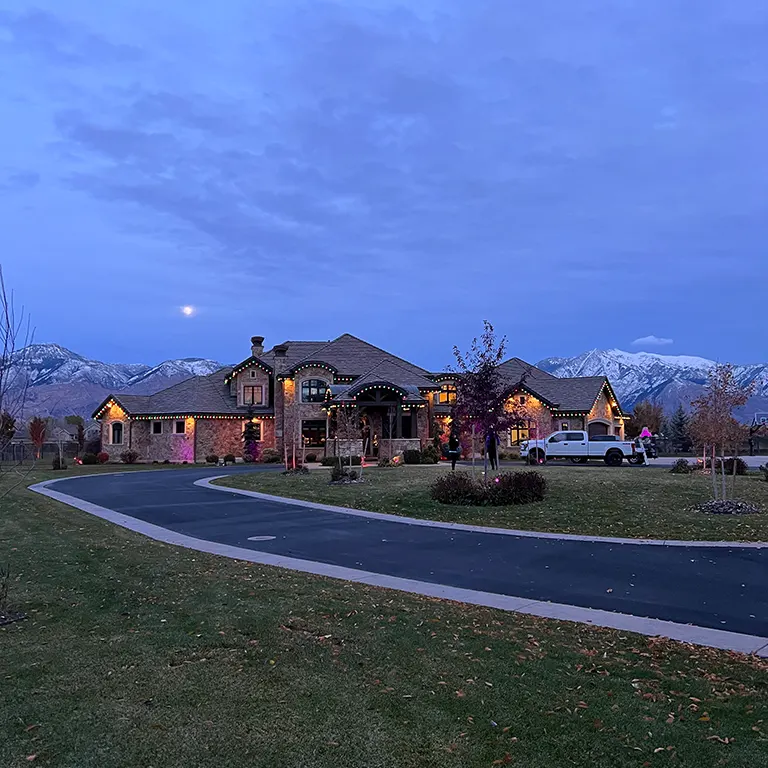 Large house with holiday lights, mountain backdrop, and a curved driveway at dusk.