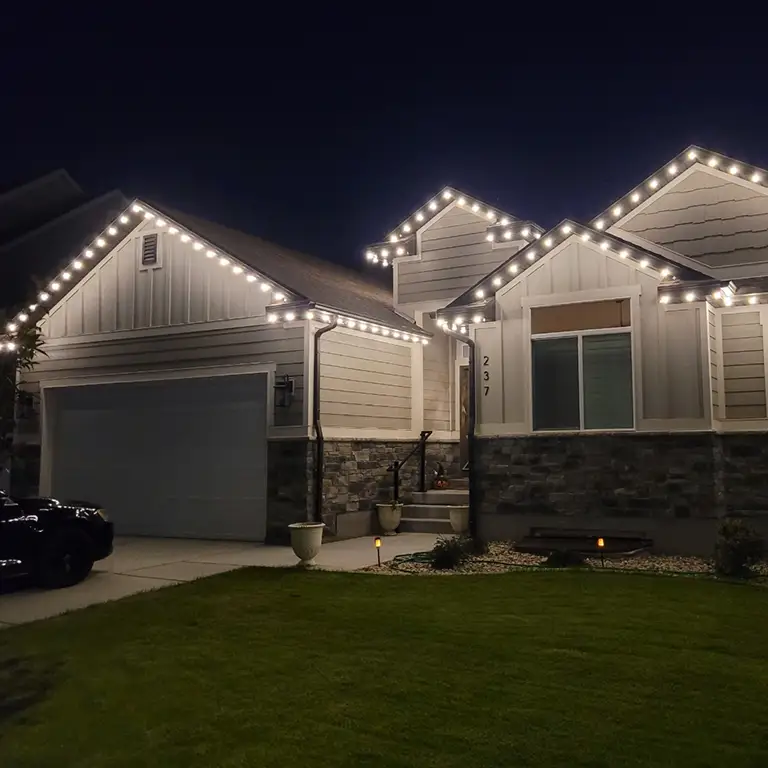 A single-story house with white trim, accented by warm white string lights outlining the roofline at night.